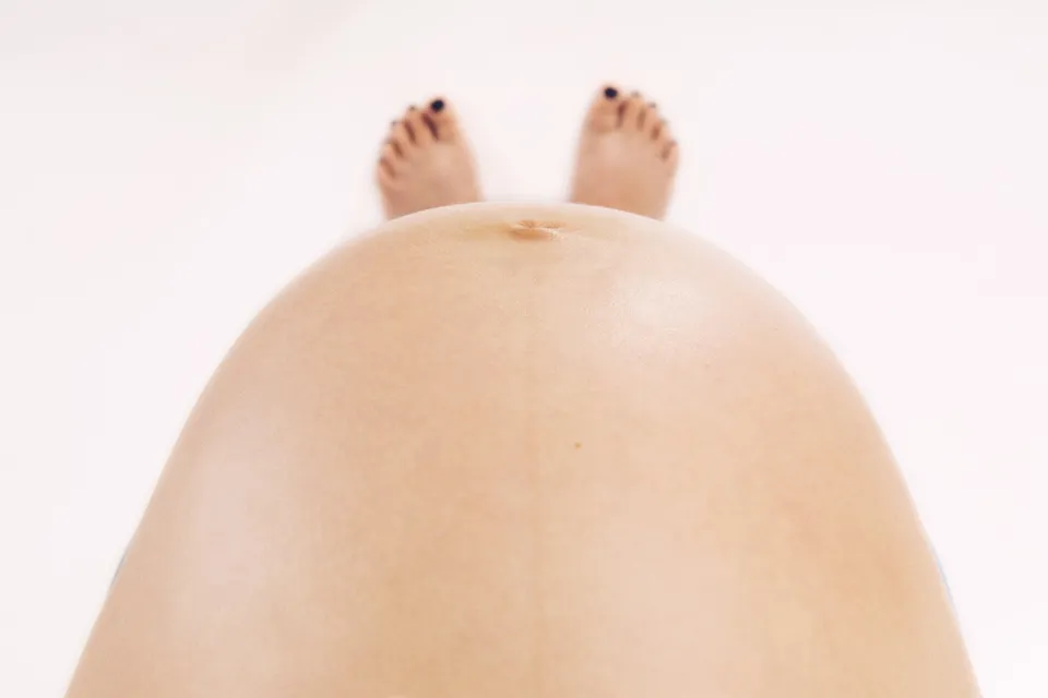 Abdomen de mujer embarazada visto desde arriba, con los pies al fondo sobre fondo claro.