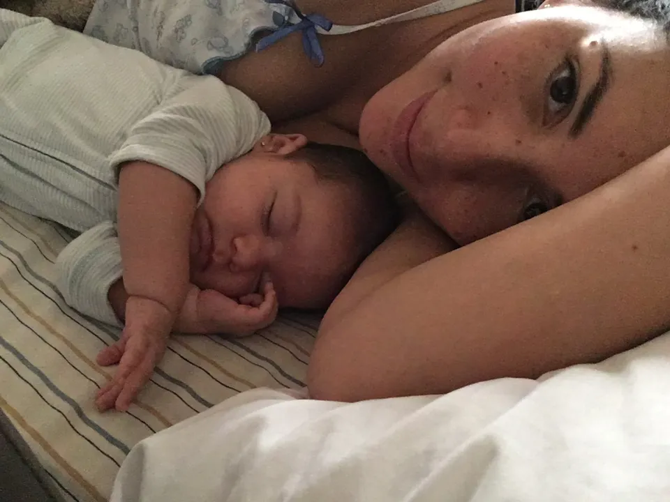 Una mujer está acostada junto a su bebé dormido, abrazándolo en una cama.