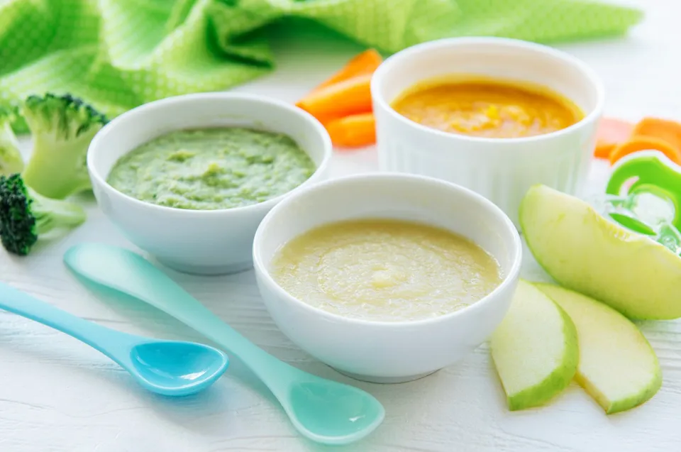 Vasitos con purés de frutas y verduras, cucharitas de colores, manzana y brócoli alrededor.