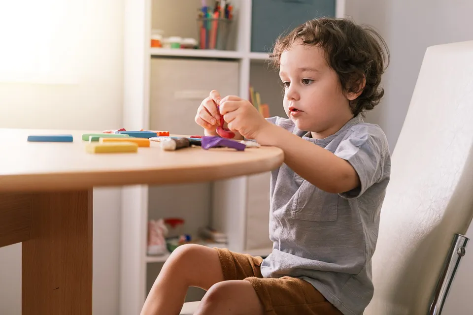 Niño pequeño sentado en una mesa jugando con plastilina de colores en casa.